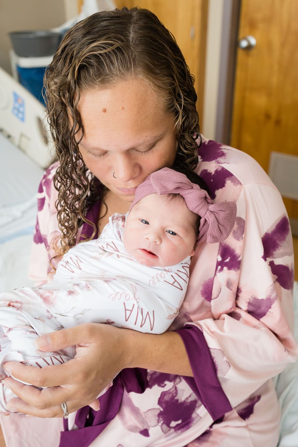 Mother in pink floral robe kissing newborn baby girl named Mia wearing pink bow in hospital room.