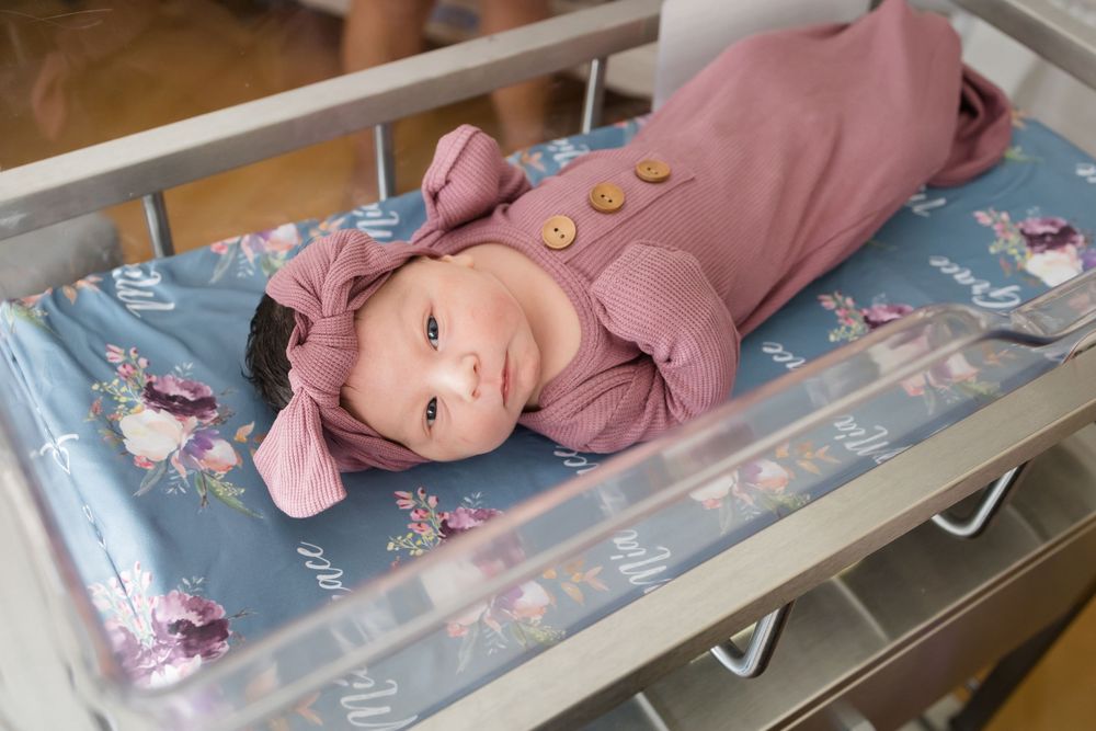 Newborn baby in pink outfit and bow headband lying in hospital bassinet on floral blanket.