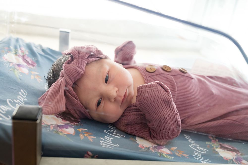 Newborn baby girl in pink romper and floral headband lying on a decorative blanket in a hospital bed.