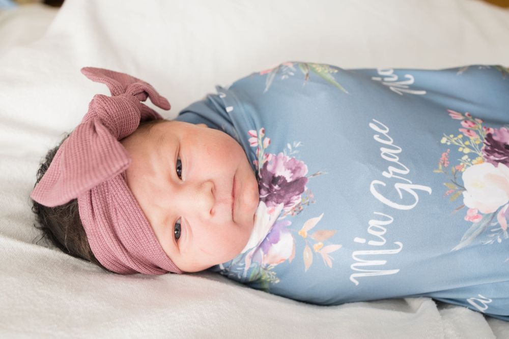 Newborn baby girl swaddled in a blue floral personalized blanket reading Mia Grace, wearing a mauve bow headband.