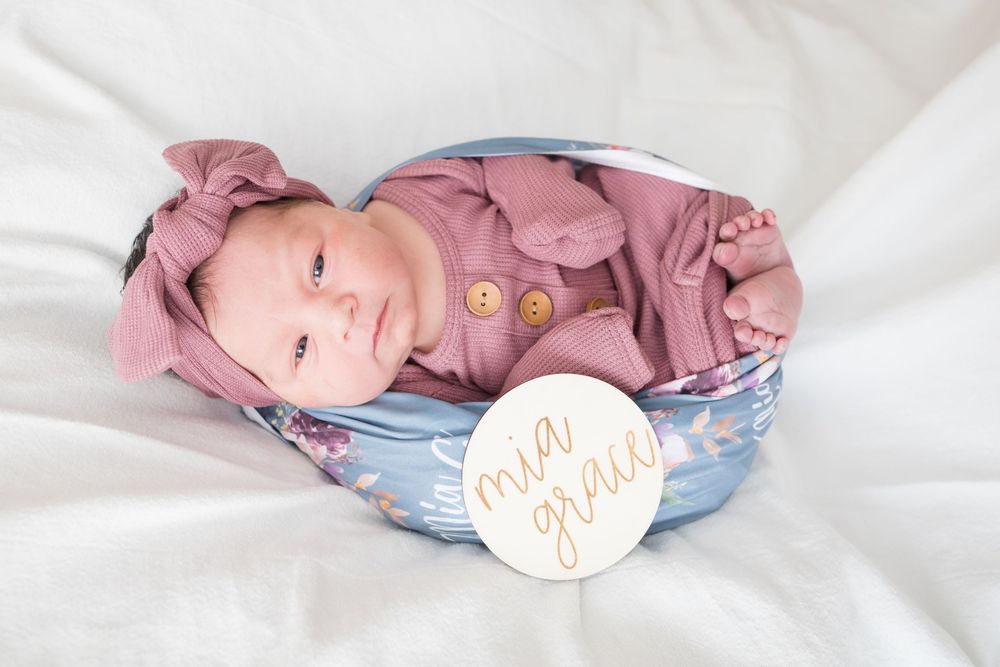 Newborn baby girl named Mia Grace in pink romper posed in bowl with floral blanket and name sign.
