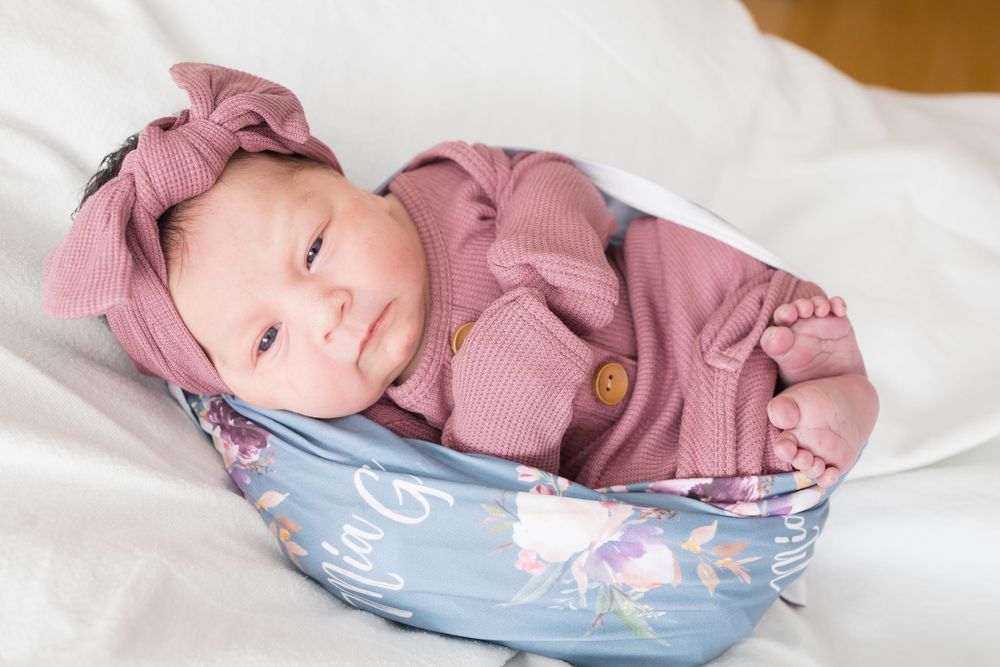 Newborn baby in pink outfit and floral headband resting in a decorative blue bowl on white fabric.