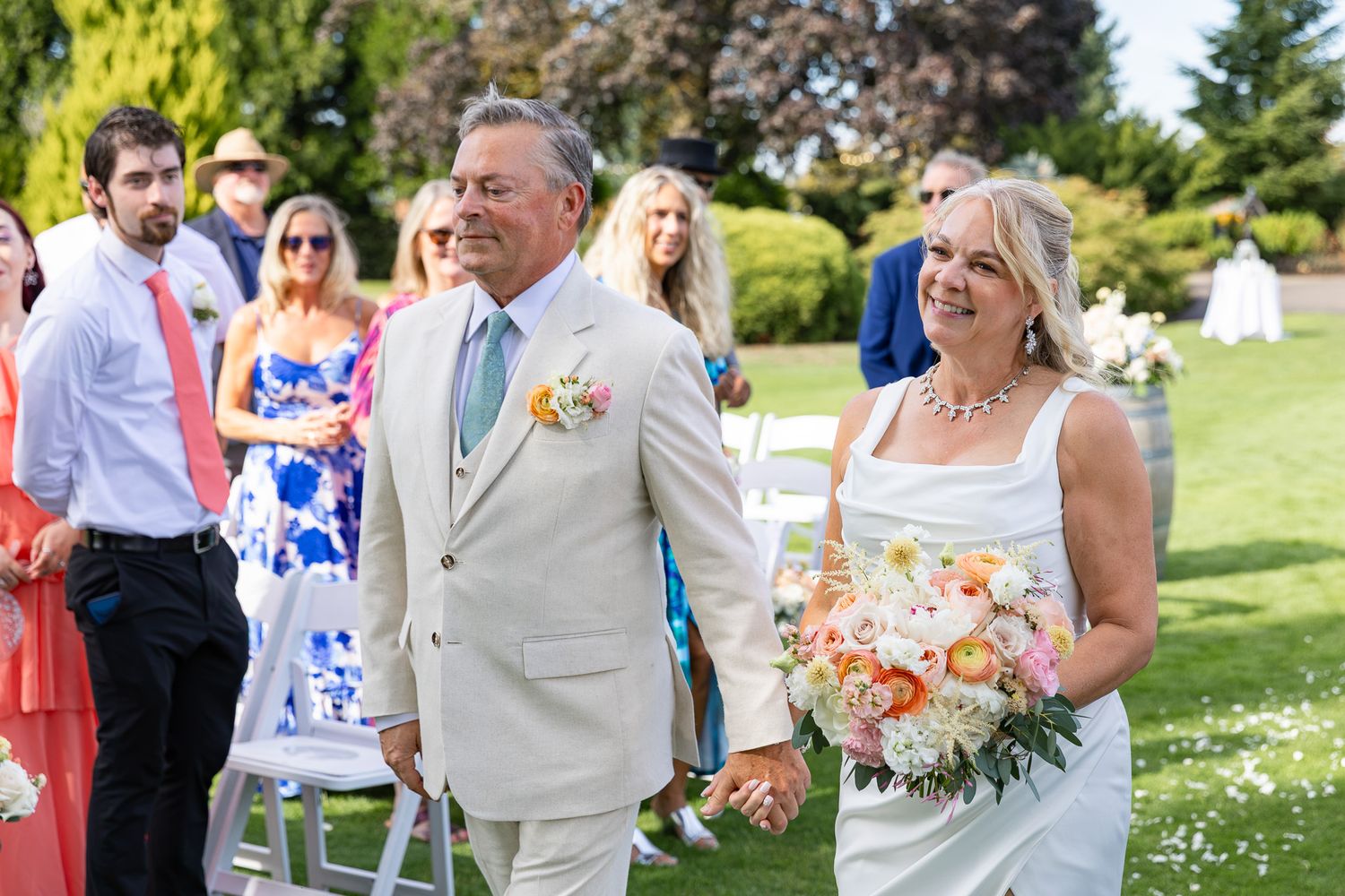 Bride and groom walk down outdoor wedding aisle, she holds colorful bouquet, he wears tan suit with floral boutonniere.