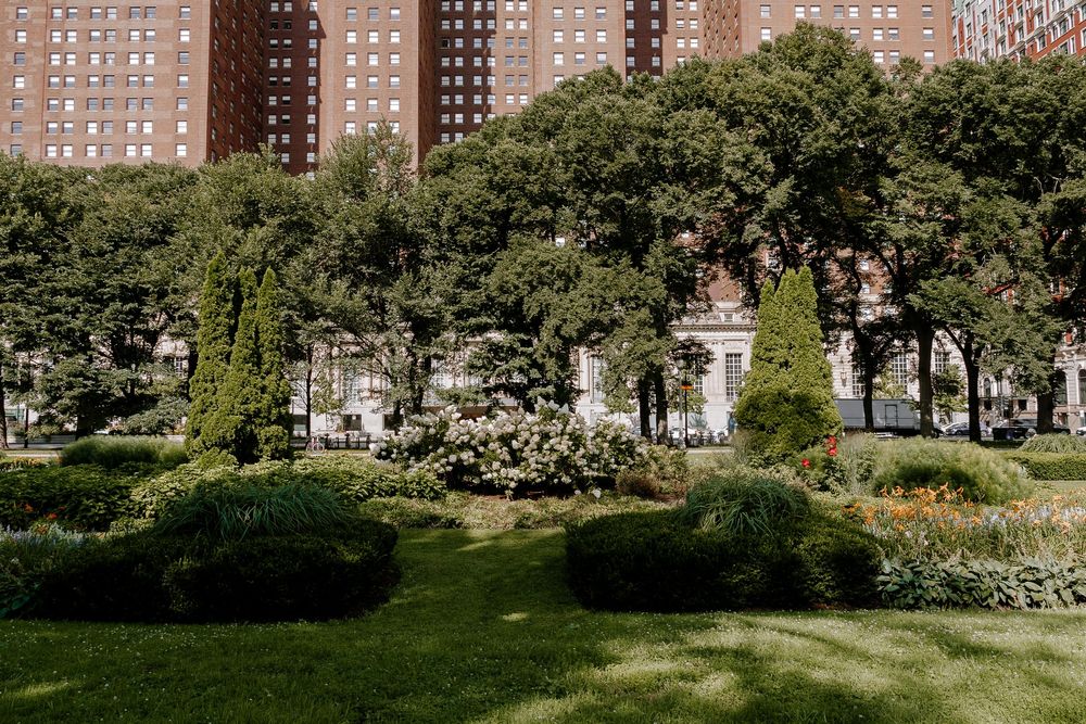 Lush urban park with manicured gardens, evergreen shrubs, and tall brick residential towers in the background on a sunny day.