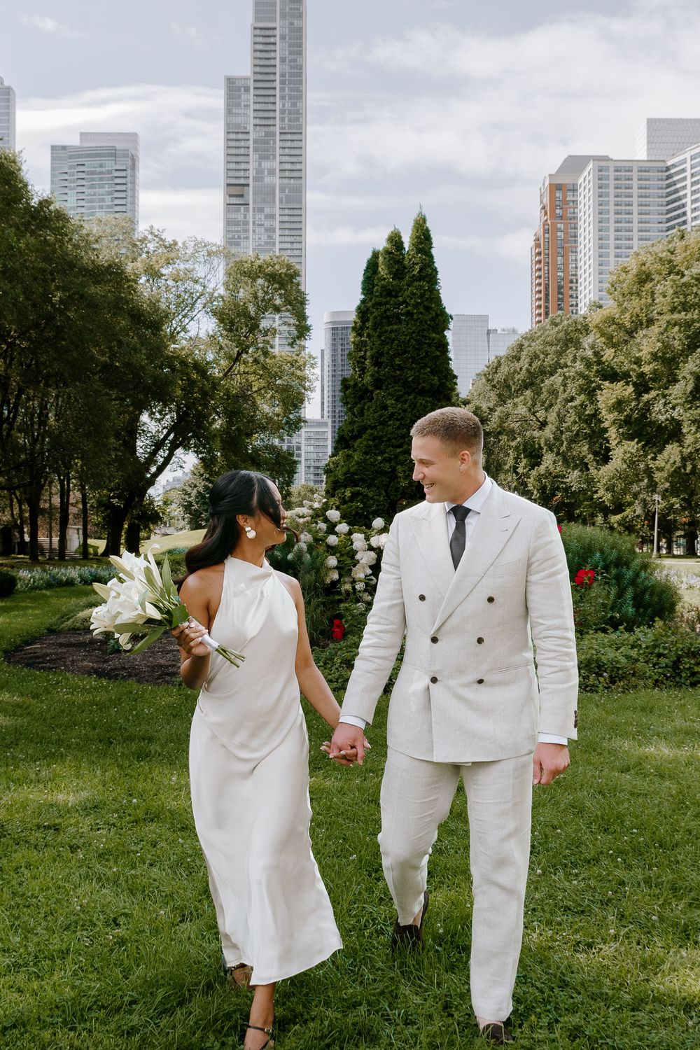 A couple in white wedding attire holds hands in a lush park with city skyscrapers in the background.