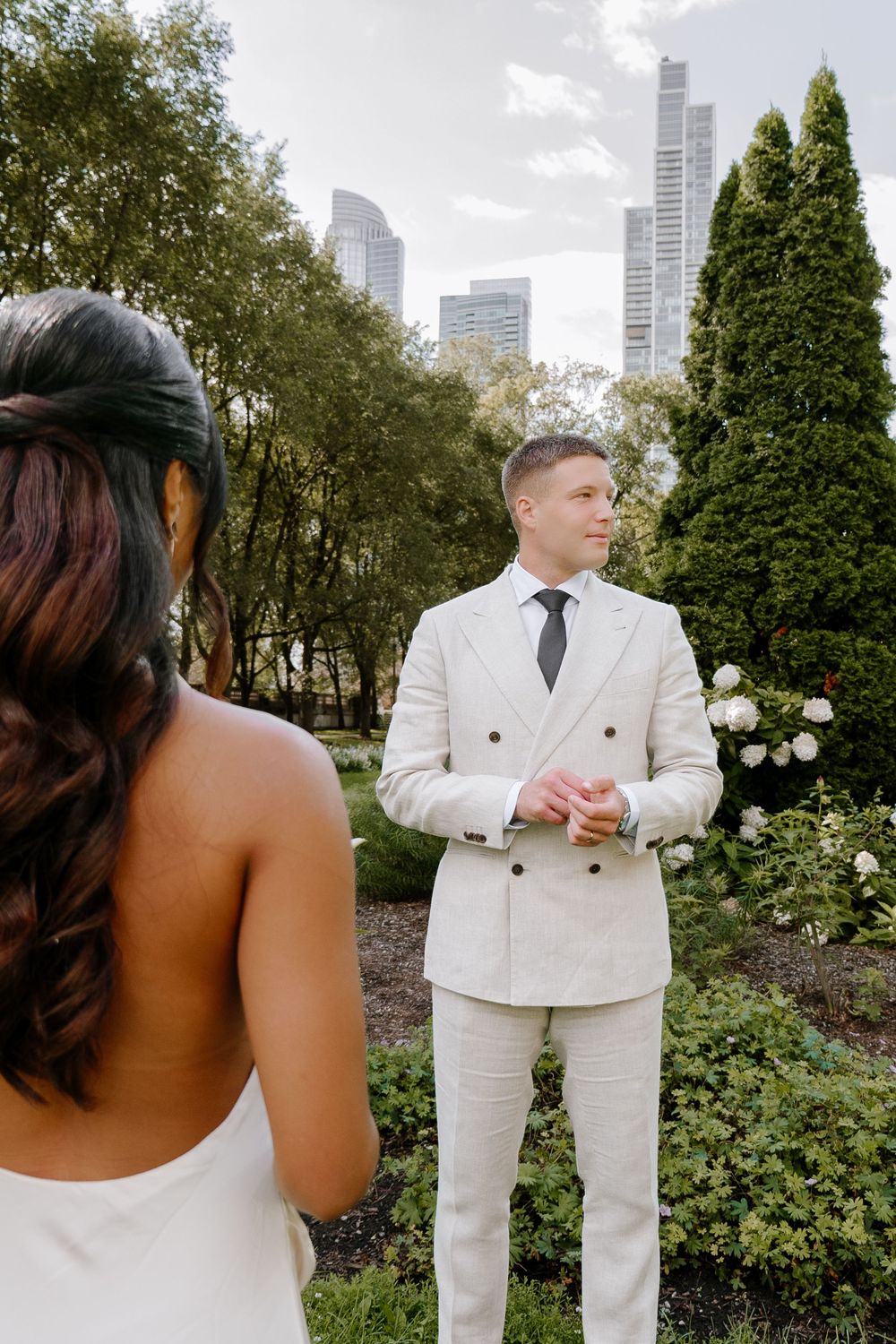 Groom in cream suit awaits bride in lush garden with city skyline backdrop.