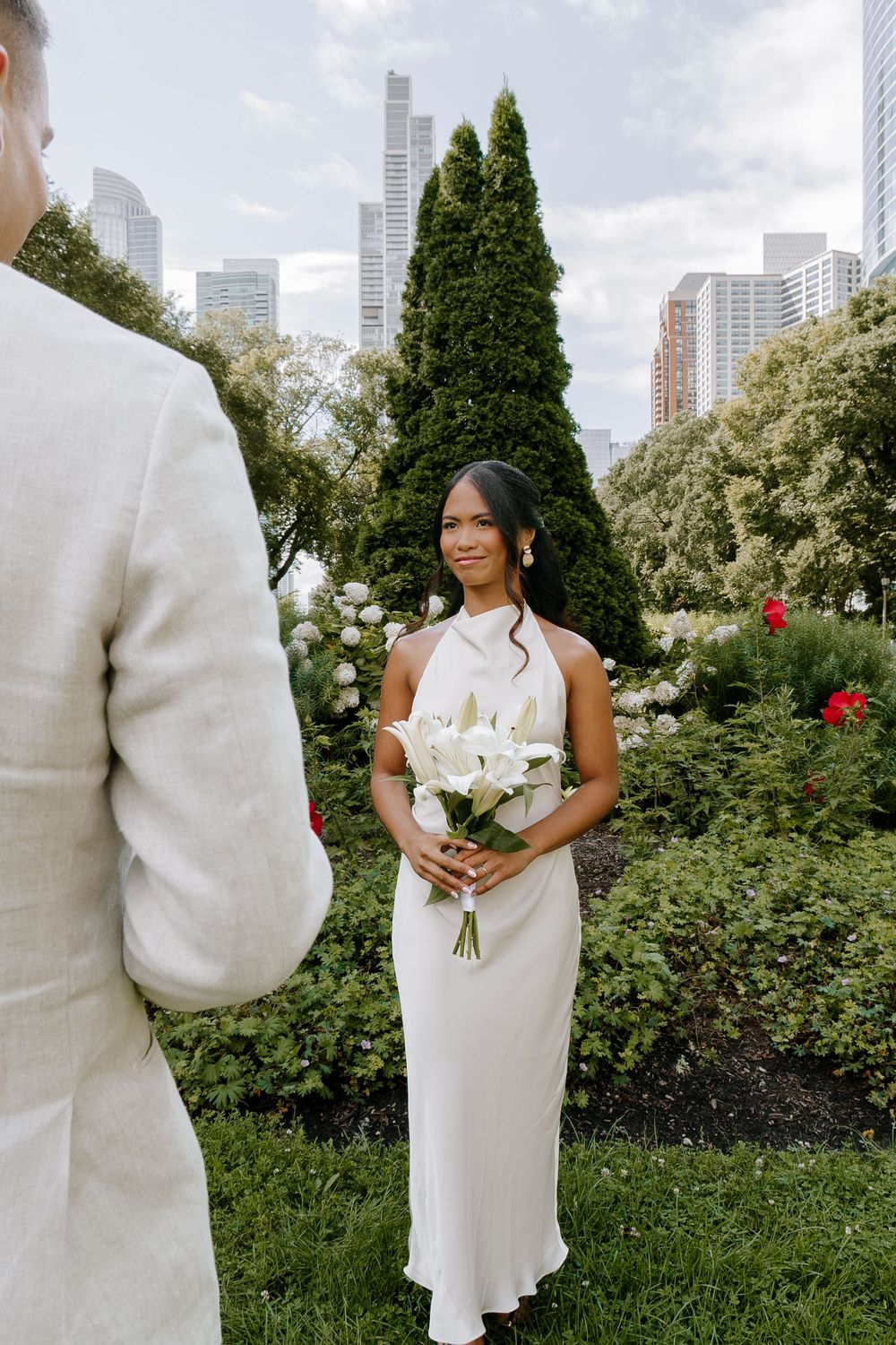 Bride in white halter gown holds calla lily bouquet in garden with city skyline backdrop.
