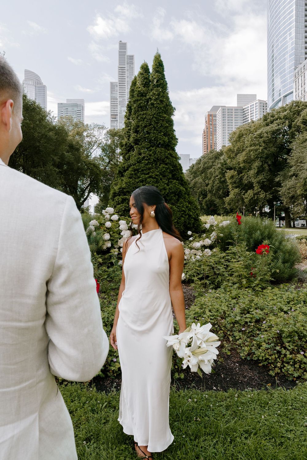 Bride in white halter gown holds lily bouquet in urban garden, groom in tan suit beside her, city skyline backdrop.