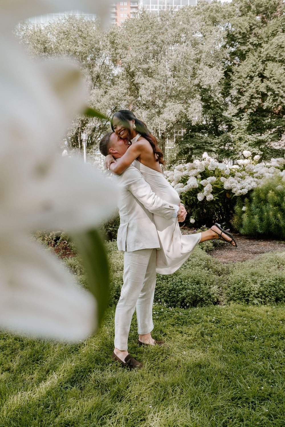 Groom in white suit lifts bride in garden, sharing romantic kiss surrounded by lush greenery and white blooms.