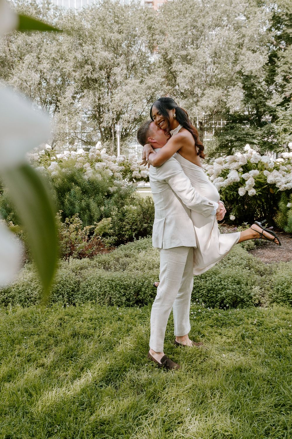 Couple in white outfits sharing a romantic lift and kiss in a lush green garden setting.