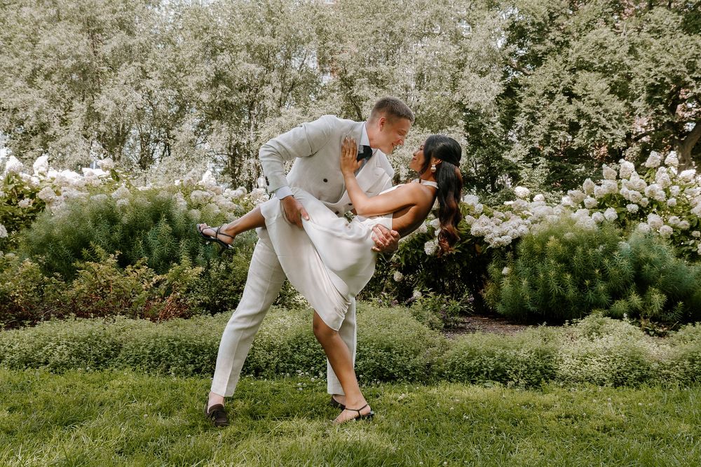 Couple sharing romantic dip kiss in lush garden with blooming white flowers, groom in white suit, outdoor wedding photo.