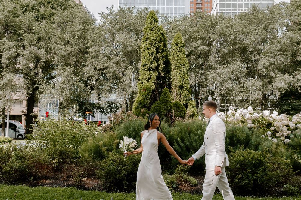 Bride and groom walk hand-in-hand through a lush garden park, bride in white gown holding bouquet.
