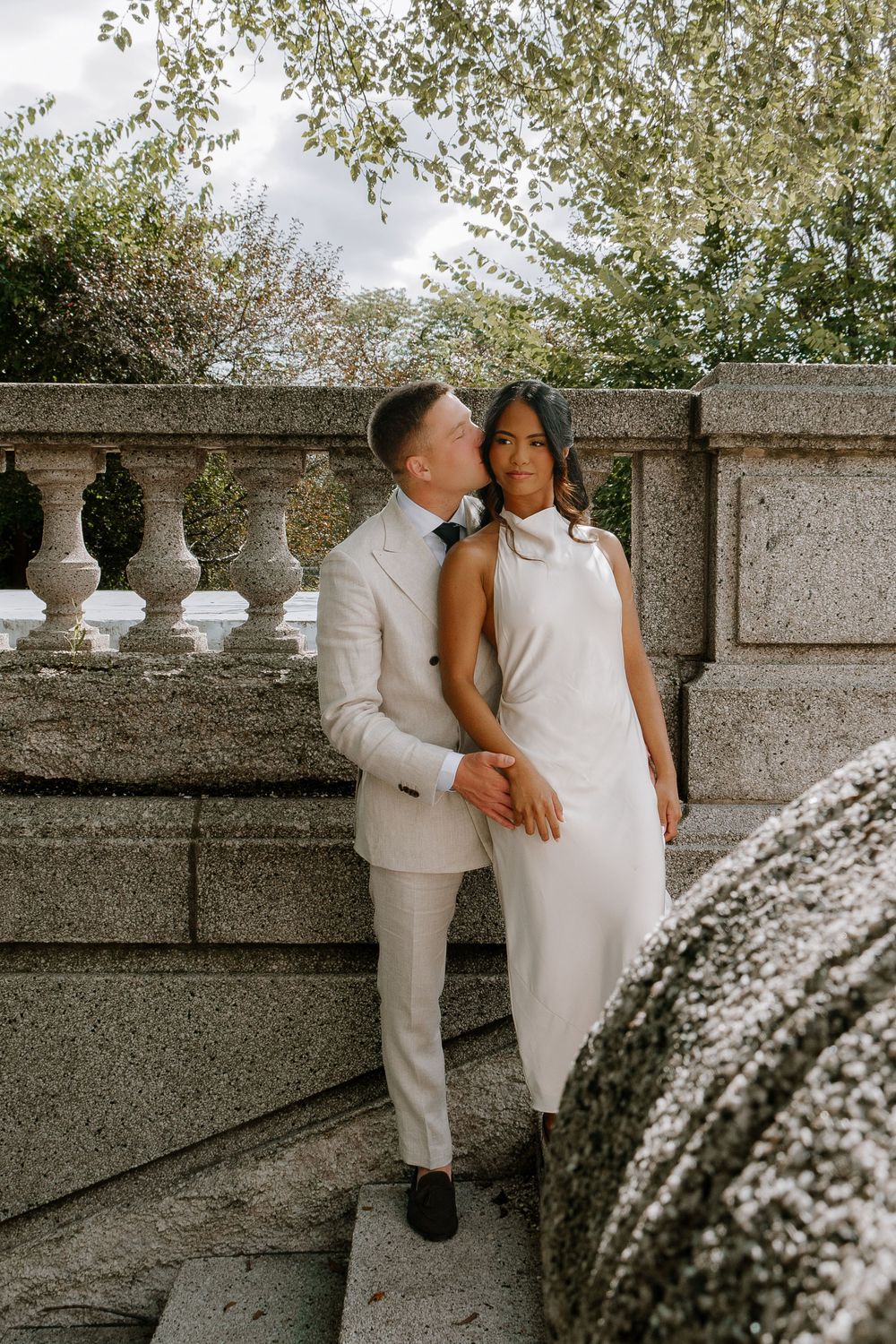 Couple in white wedding attire poses on stone steps with balustrade, groom kissing bride's cheek outdoors.