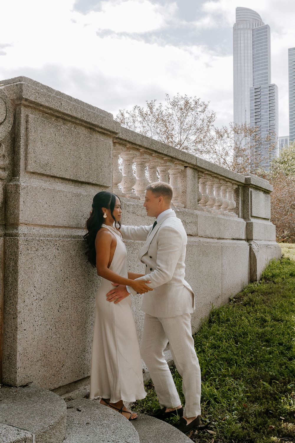 Couple in white wedding attire posing by a stone balustrade with city skyscrapers in the background.