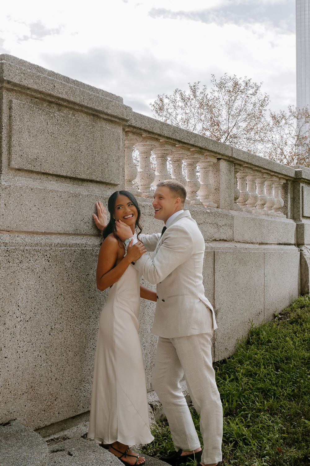 Couple in white wedding attire laughing against a stone balustrade outdoors.