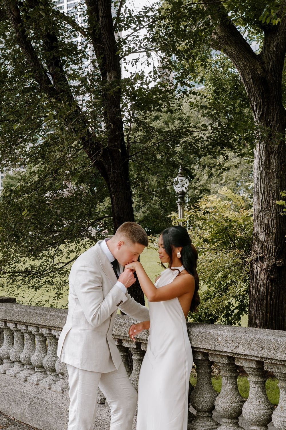 Groom in white suit kisses bride's hand on stone balcony surrounded by lush green trees in a romantic outdoor wedding portrait.