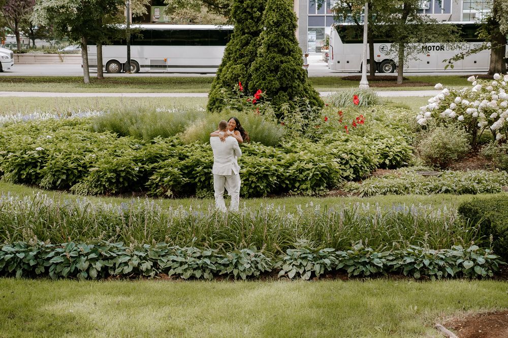 Couple embracing in a lush garden with green hedges, colorful flowers, and trees on a cloudy day.