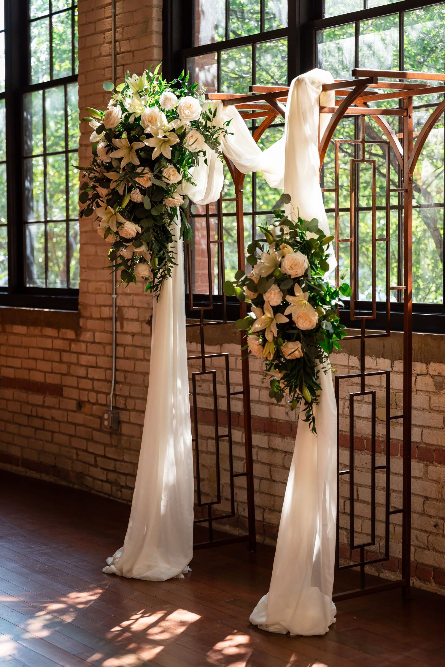 Elegant wedding arch with white draping and blush roses, lilies, and greenery arrangements in a rustic brick venue.