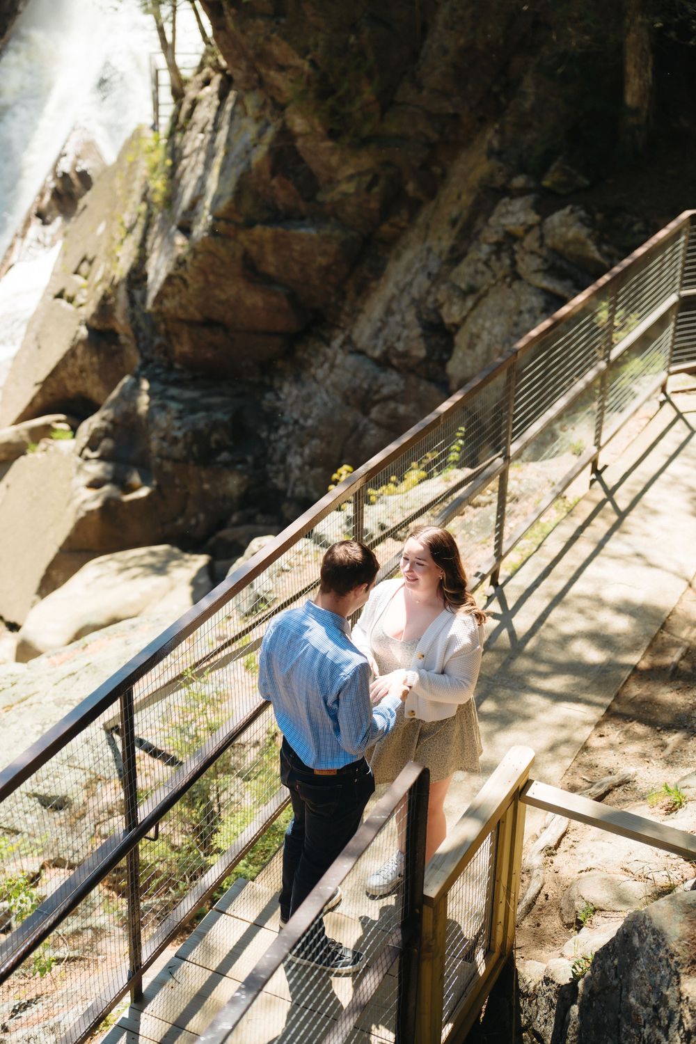 Couple holding hands on a metal bridge staircase surrounded by rocky cliffs in a scenic outdoor setting.