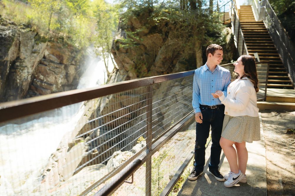 Couple standing on a scenic walkway beside a waterfall, smiling at each other near metal railings and stone steps.