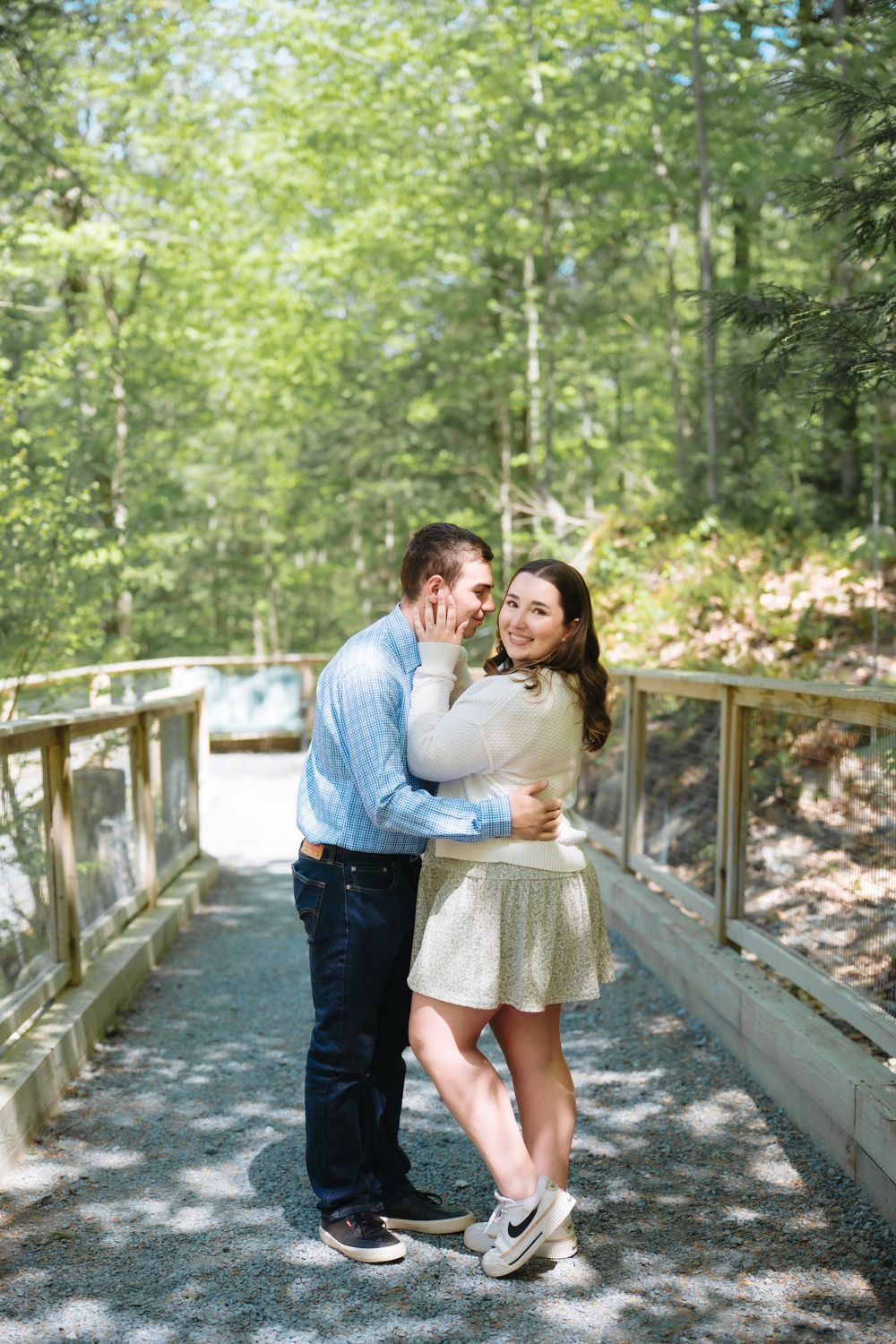 Couple embracing on a wooden bridge surrounded by lush green trees, woman smiling in white top and gold skirt.