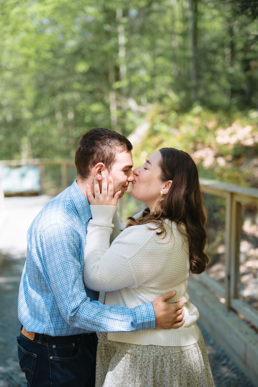 A couple shares a romantic kiss outdoors on a wooden bridge surrounded by lush green forest trees.