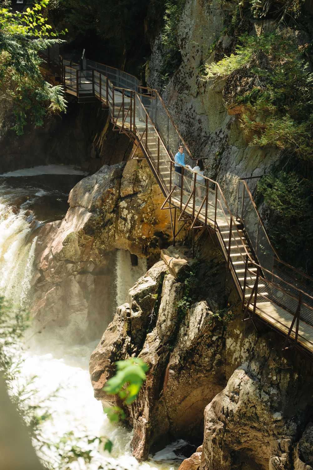 Person sitting on metal staircase built into rocky cliff face beside waterfall surrounded by lush green forest.