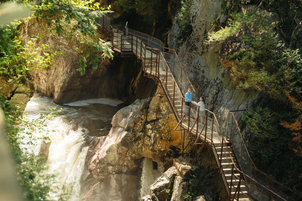 Hiker on a metal walkway bridge through a lush forest gorge with a rushing waterfall below.