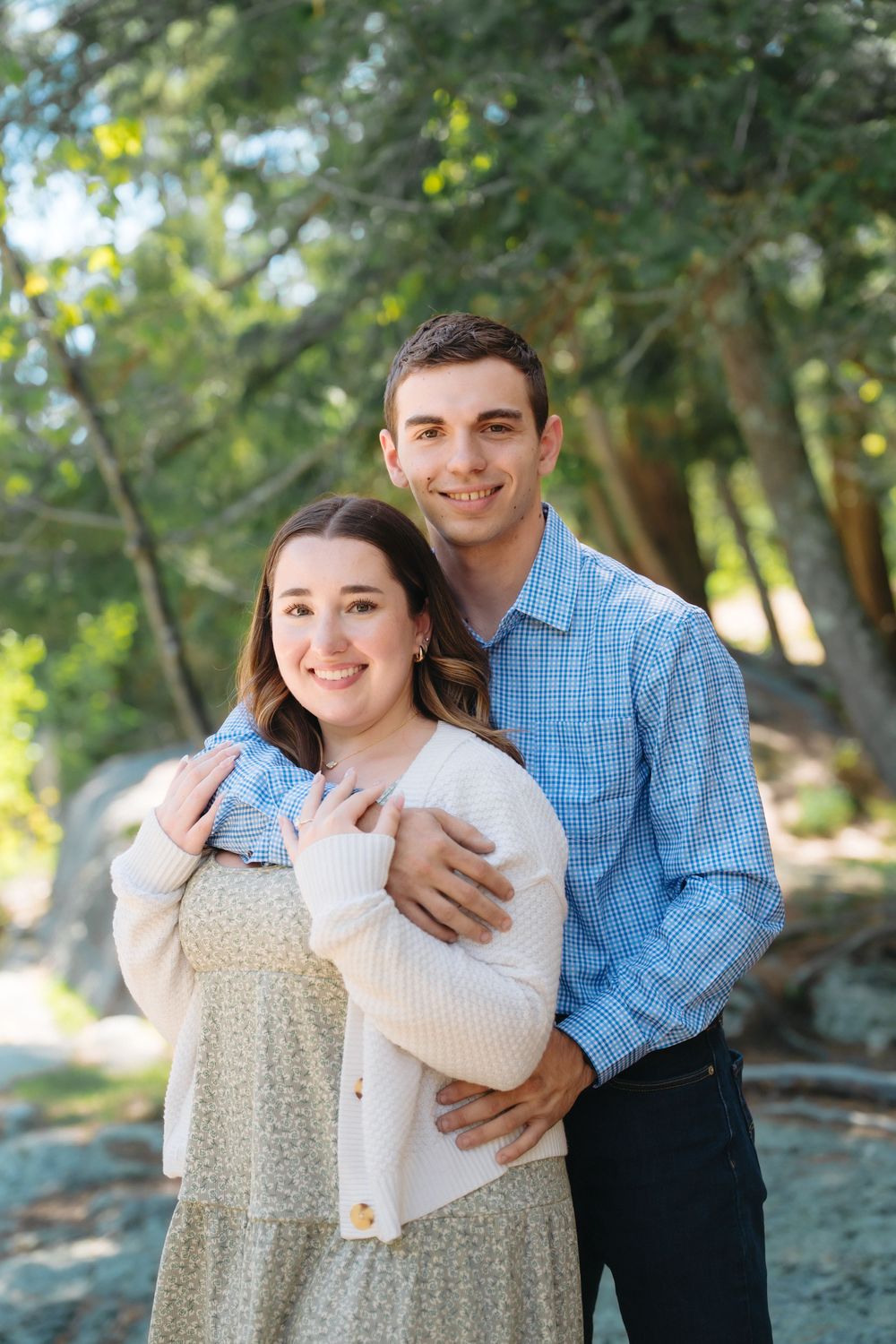 A smiling couple poses outdoors among green trees, the man embracing the woman from behind in casual attire.