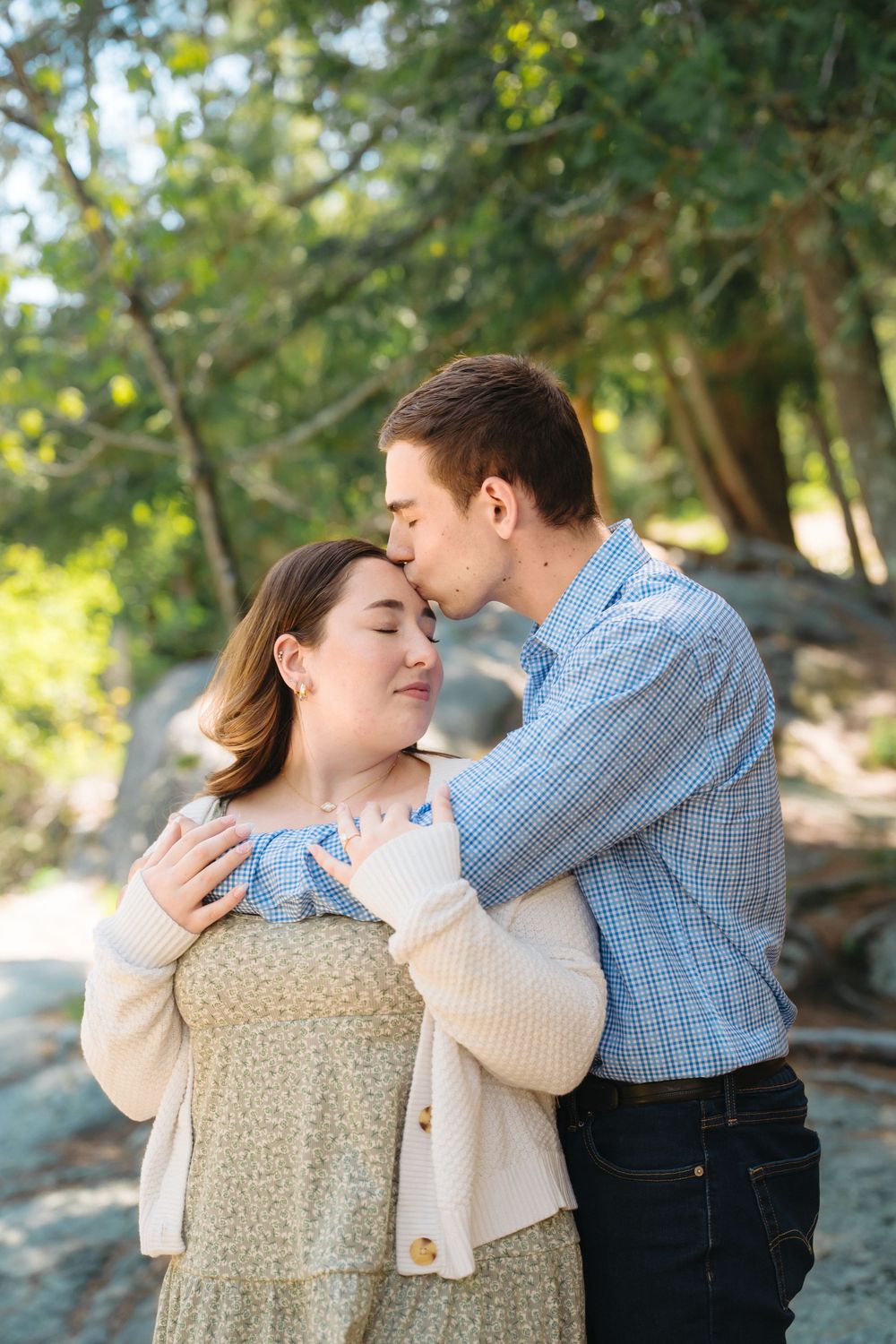 A couple shares a tender moment outdoors, man kissing woman's temple while embracing her among lush green trees.