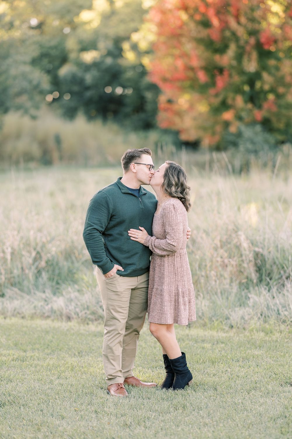 Couple sharing a kiss in a field with vibrant autumn foliage backdrop during an outdoor engagement session.