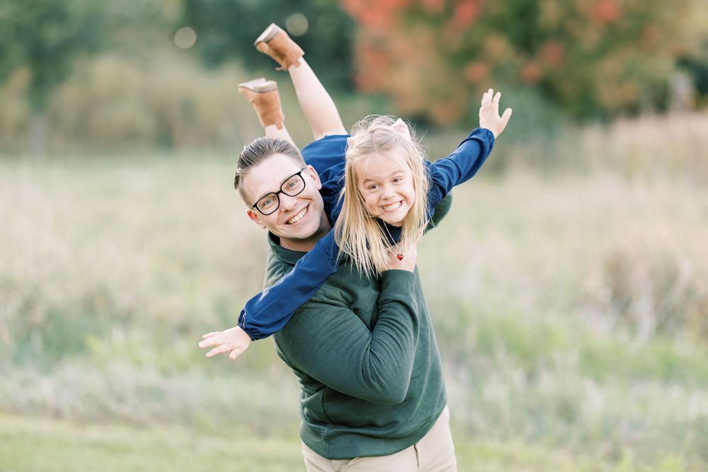 A woman gives a joyful girl a piggyback ride outdoors, both smiling with arms spread wide in a green field.