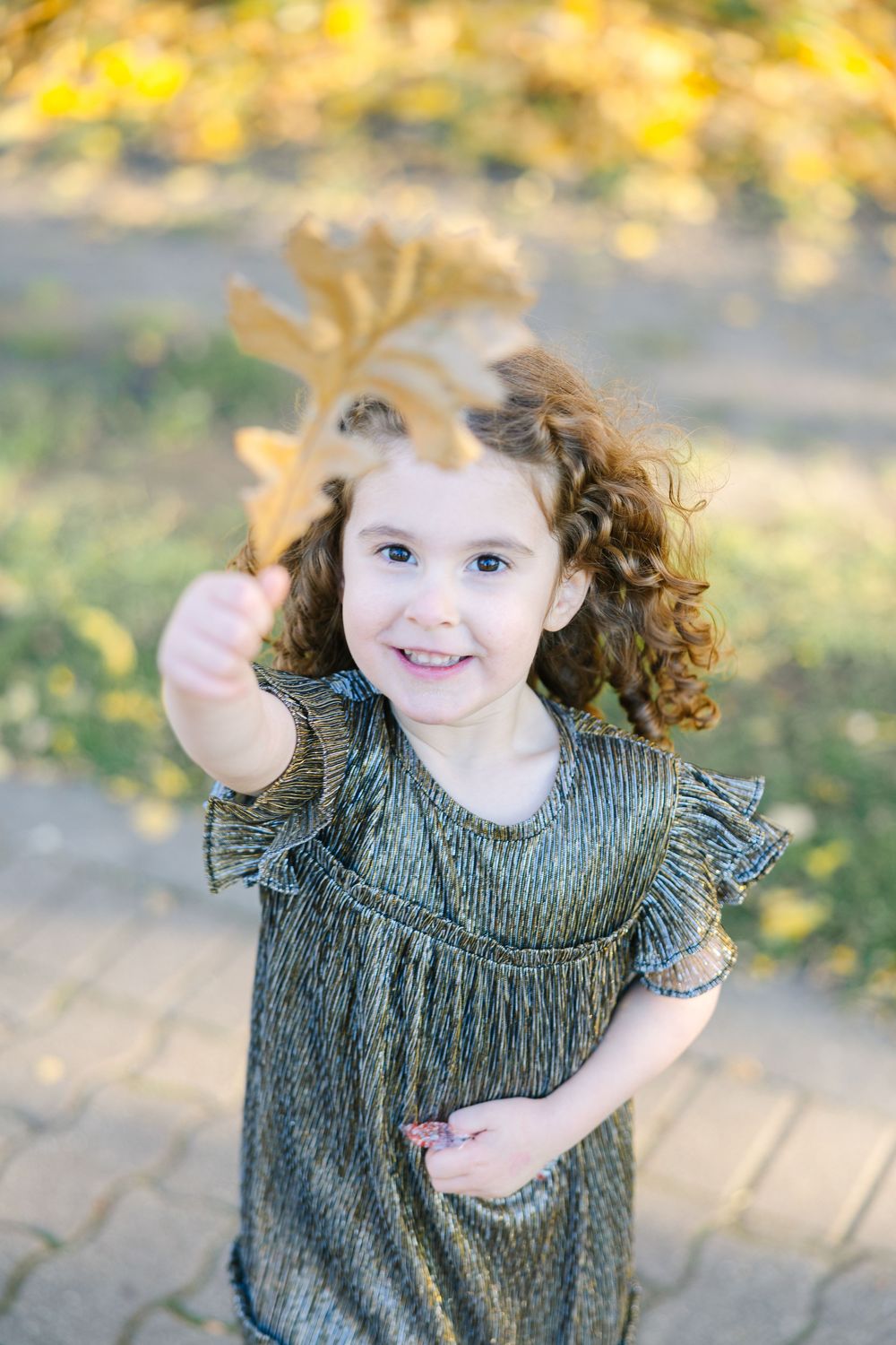 Smiling curly-haired girl in sparkly dress holding a golden leaf in an autumn park.