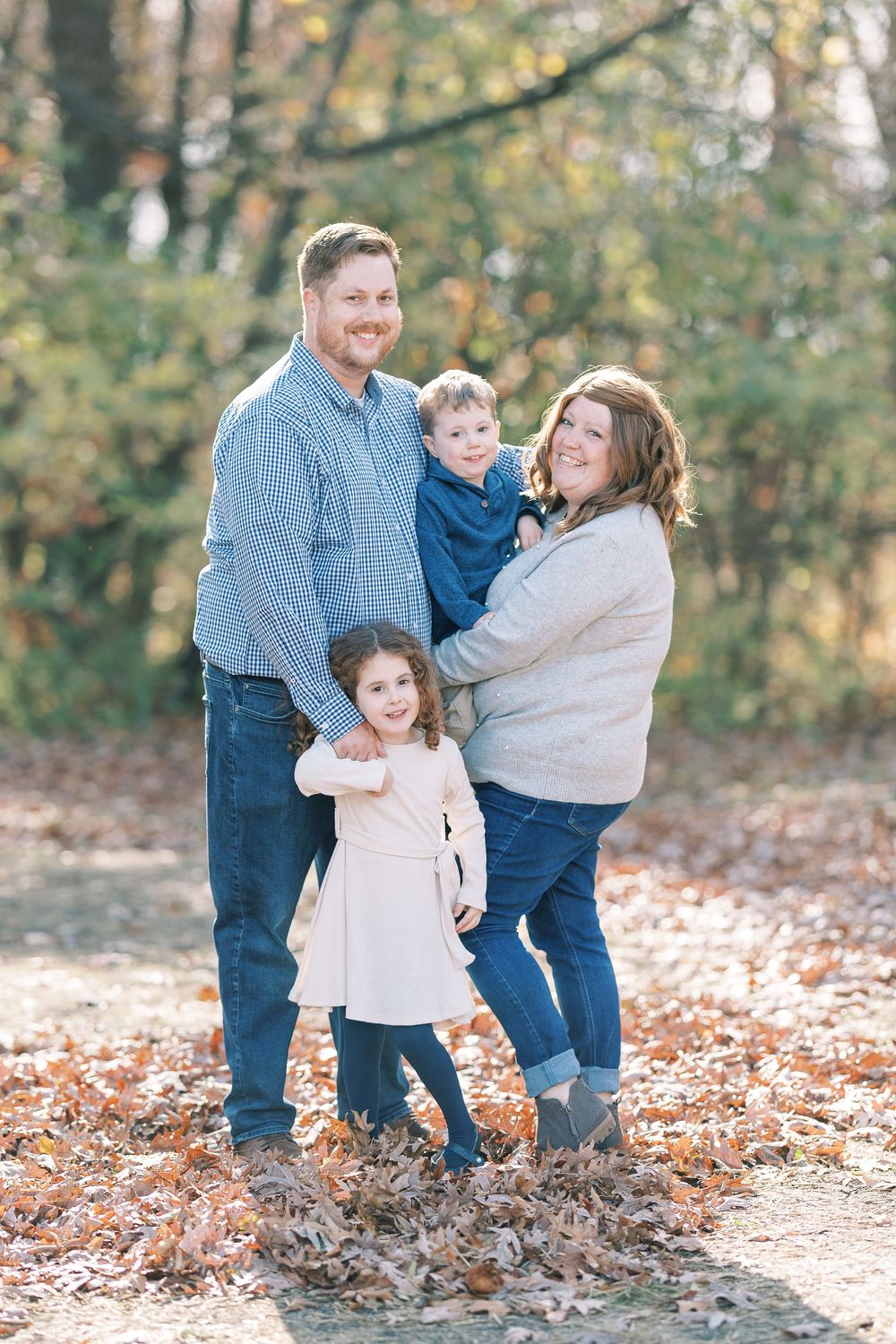 Family of four smiling outdoors in autumn leaves, parents holding young children in casual blue and neutral outfits.