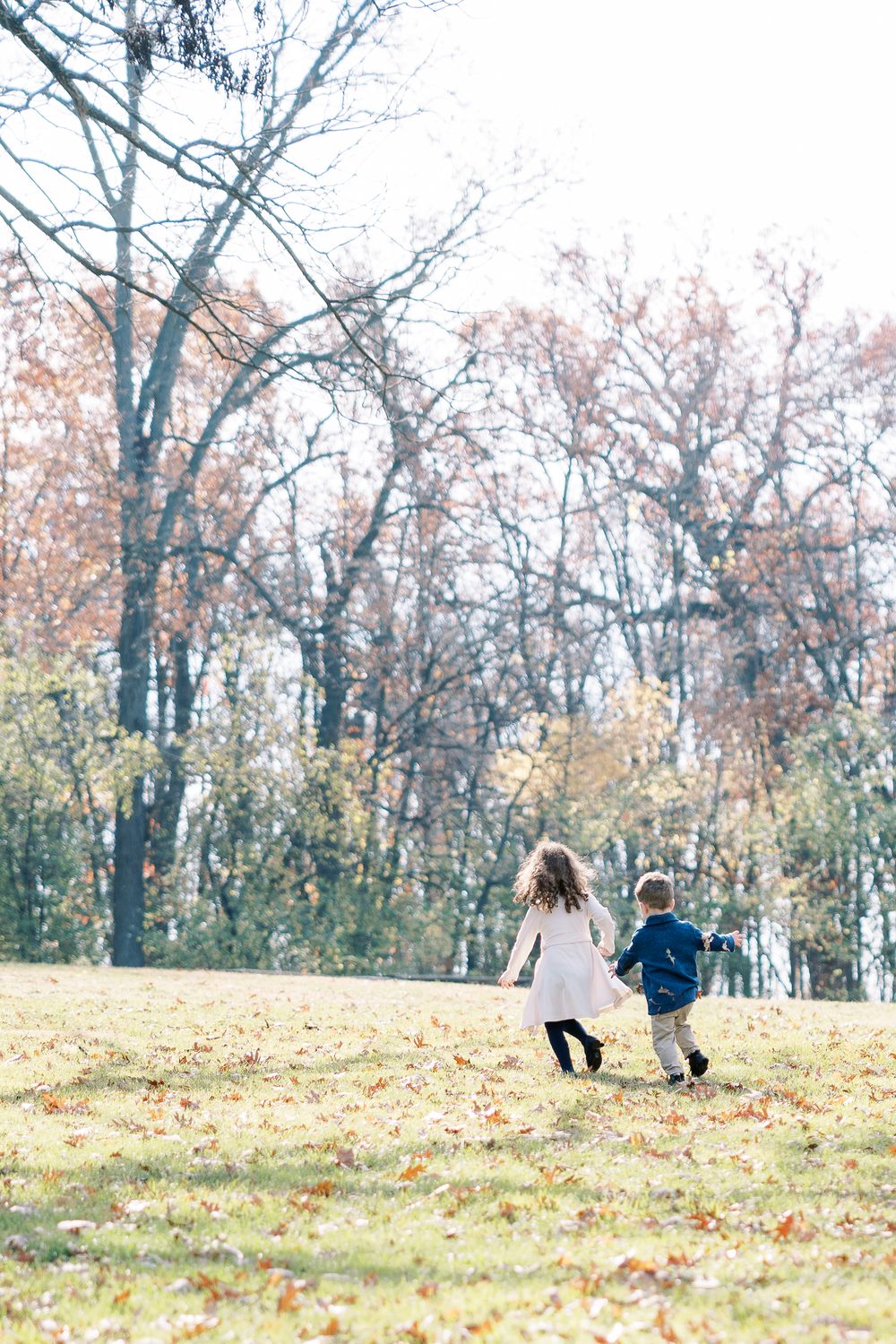 Two young children holding hands and running through an autumn park with fallen leaves and bare trees.