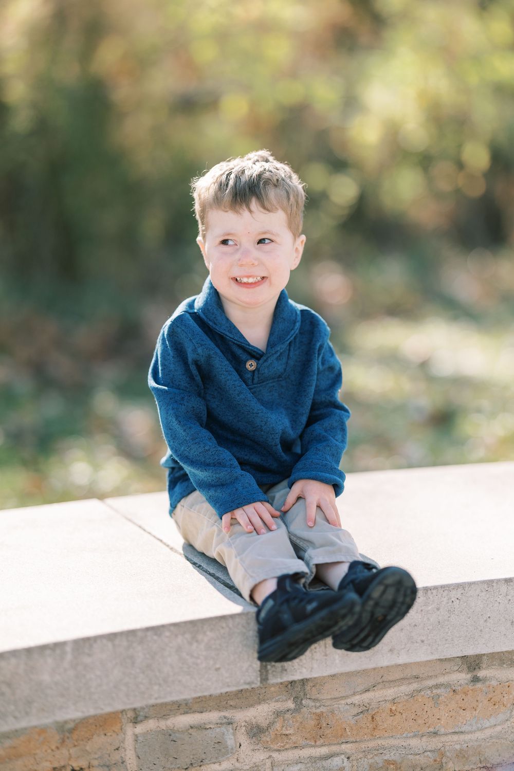 Smiling young boy in blue sweater sitting on stone wall outdoors with autumn foliage background.
