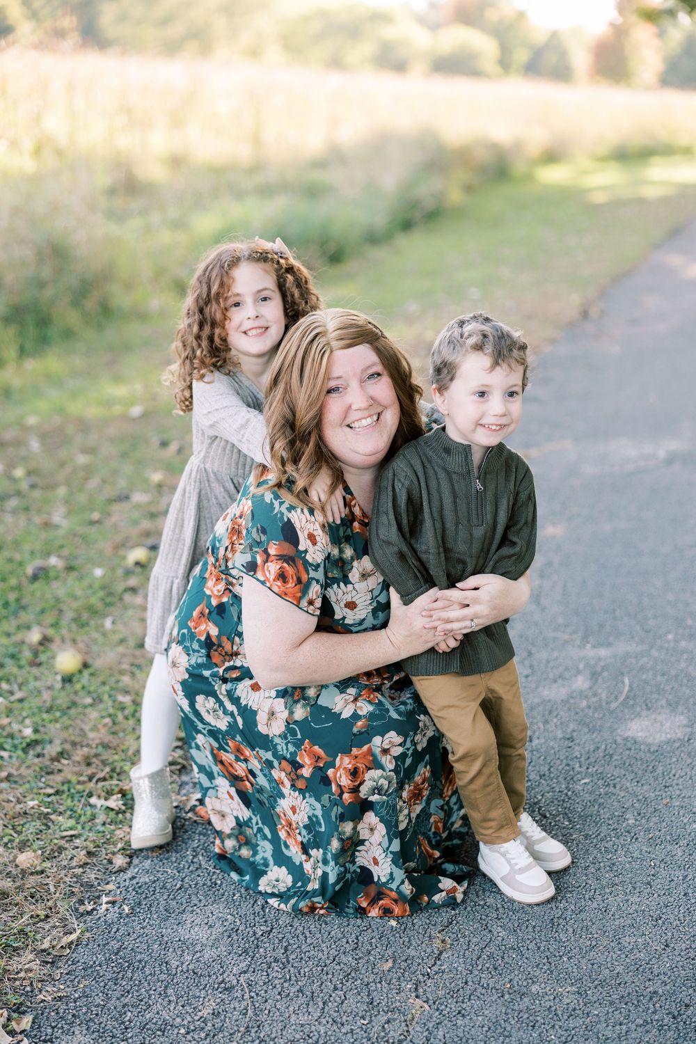 A smiling woman in a floral dress crouches on a path with two young children hugging her in a warm outdoor setting.