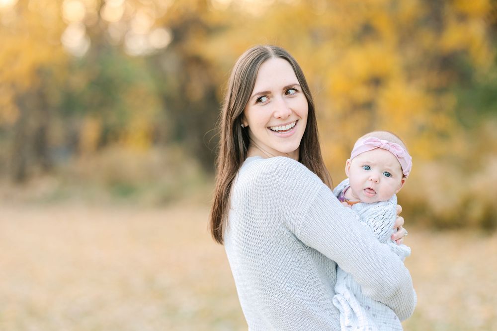 A smiling mother holding her baby girl in a pink headband outdoors among golden autumn trees.