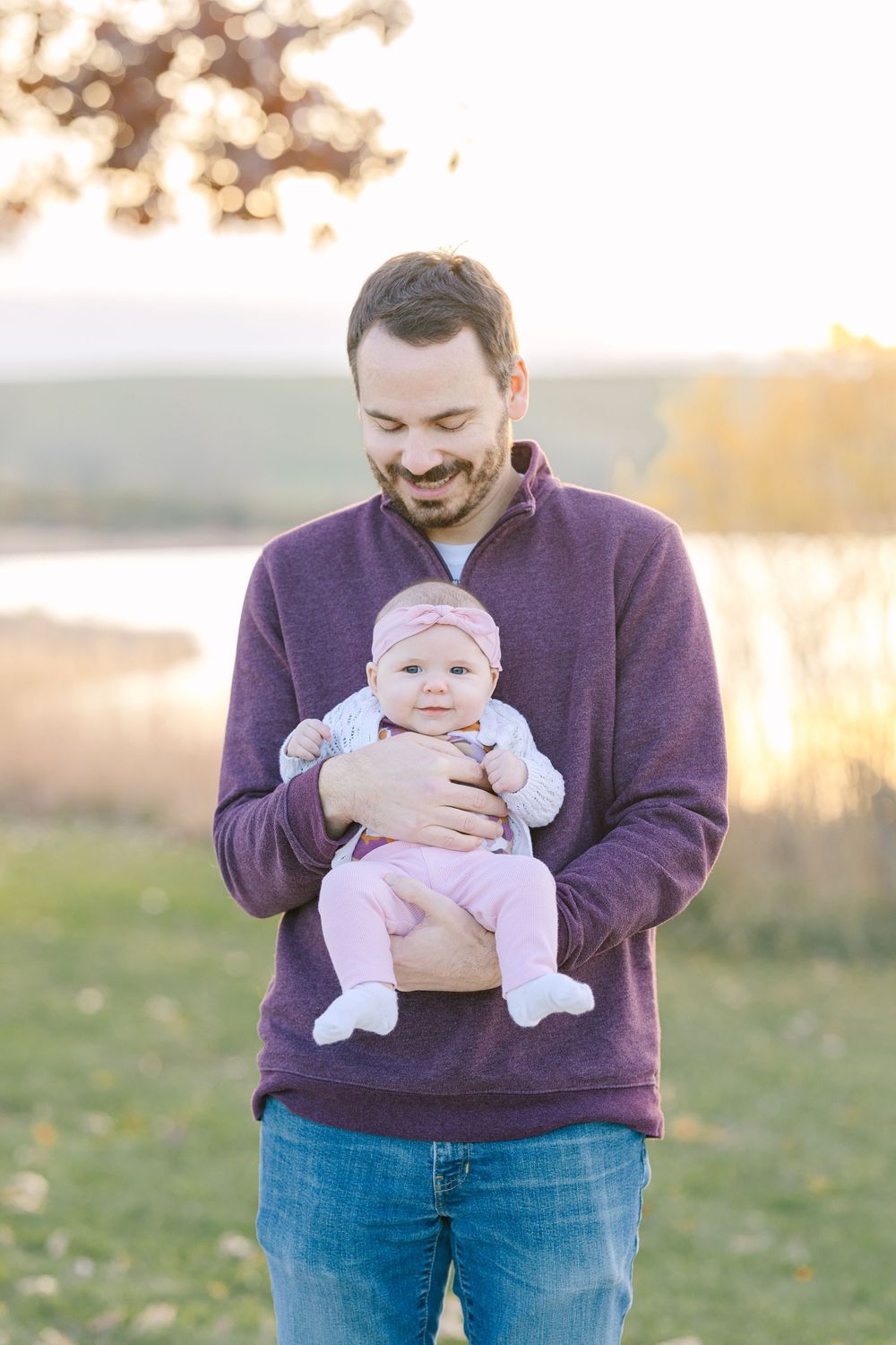 A smiling father in a purple sweater holds his baby girl outdoors near a lake during golden hour.