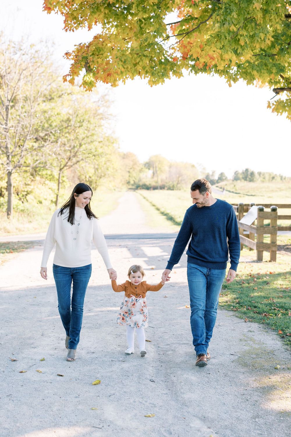 A young family walks hand-in-hand down a sunny autumn path, parents swinging their toddler between them.