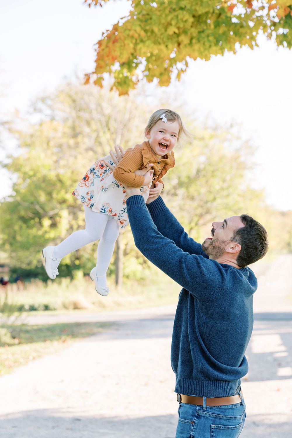 A father joyfully lifts his laughing toddler daughter into the air outdoors during a sunny autumn day.