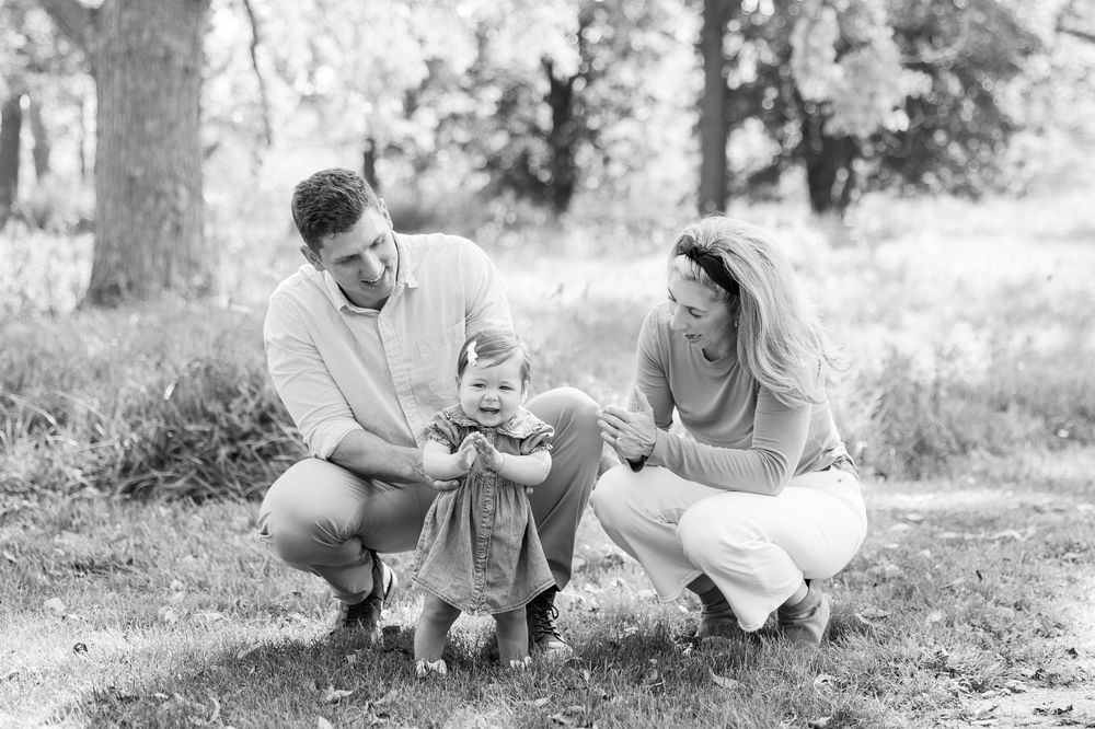 Black and white family photo of parents crouching with their baby girl standing between them outdoors in a park.