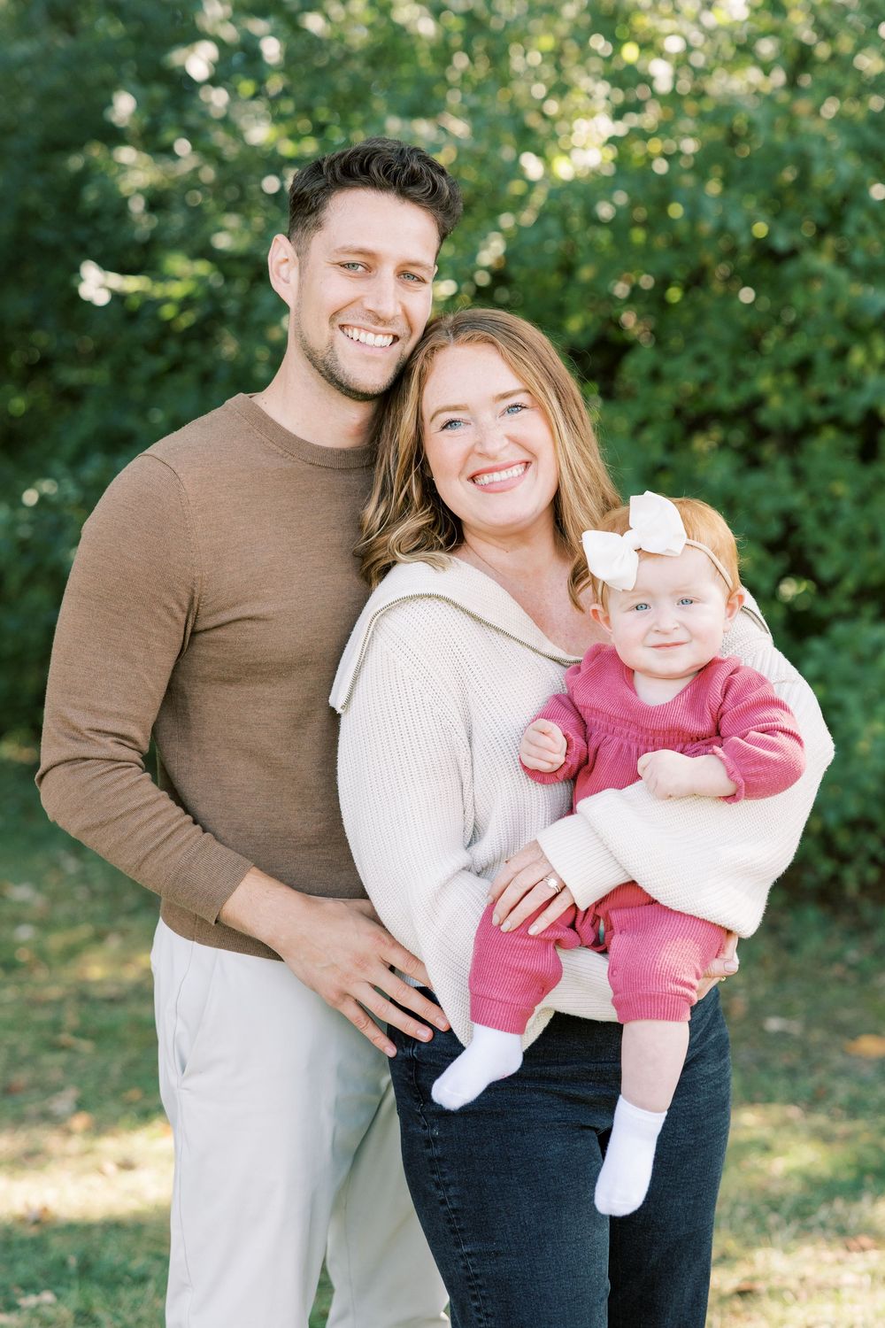Smiling couple holding baby girl in pink outfit outdoors with green trees in background.