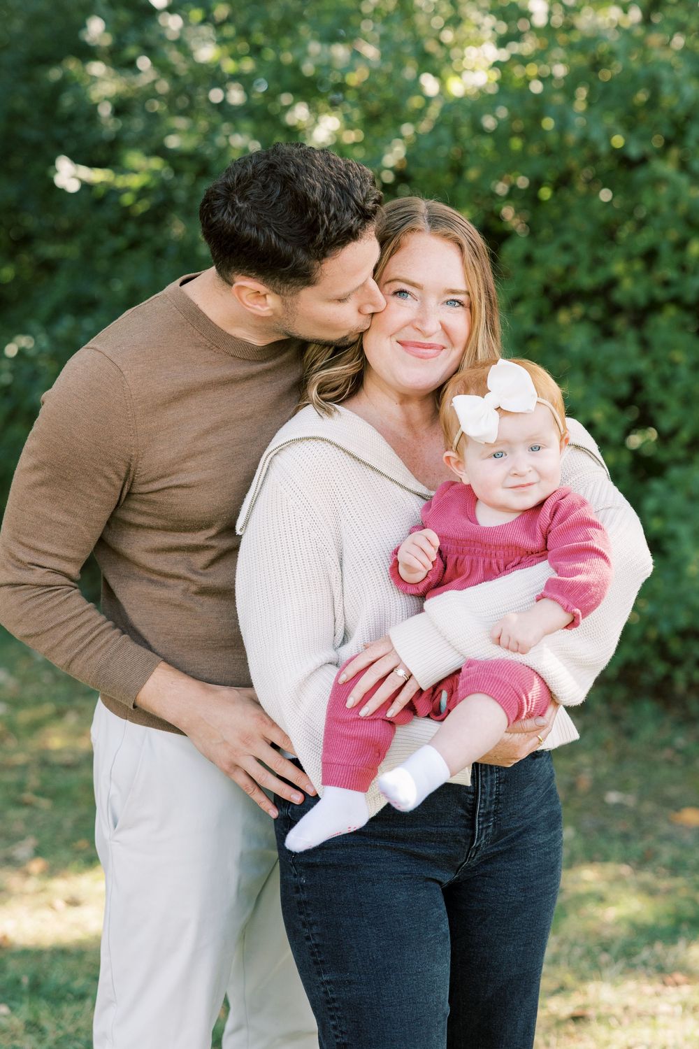 A happy young family with a man kissing a smiling woman on the cheek as she holds their baby girl outdoors.