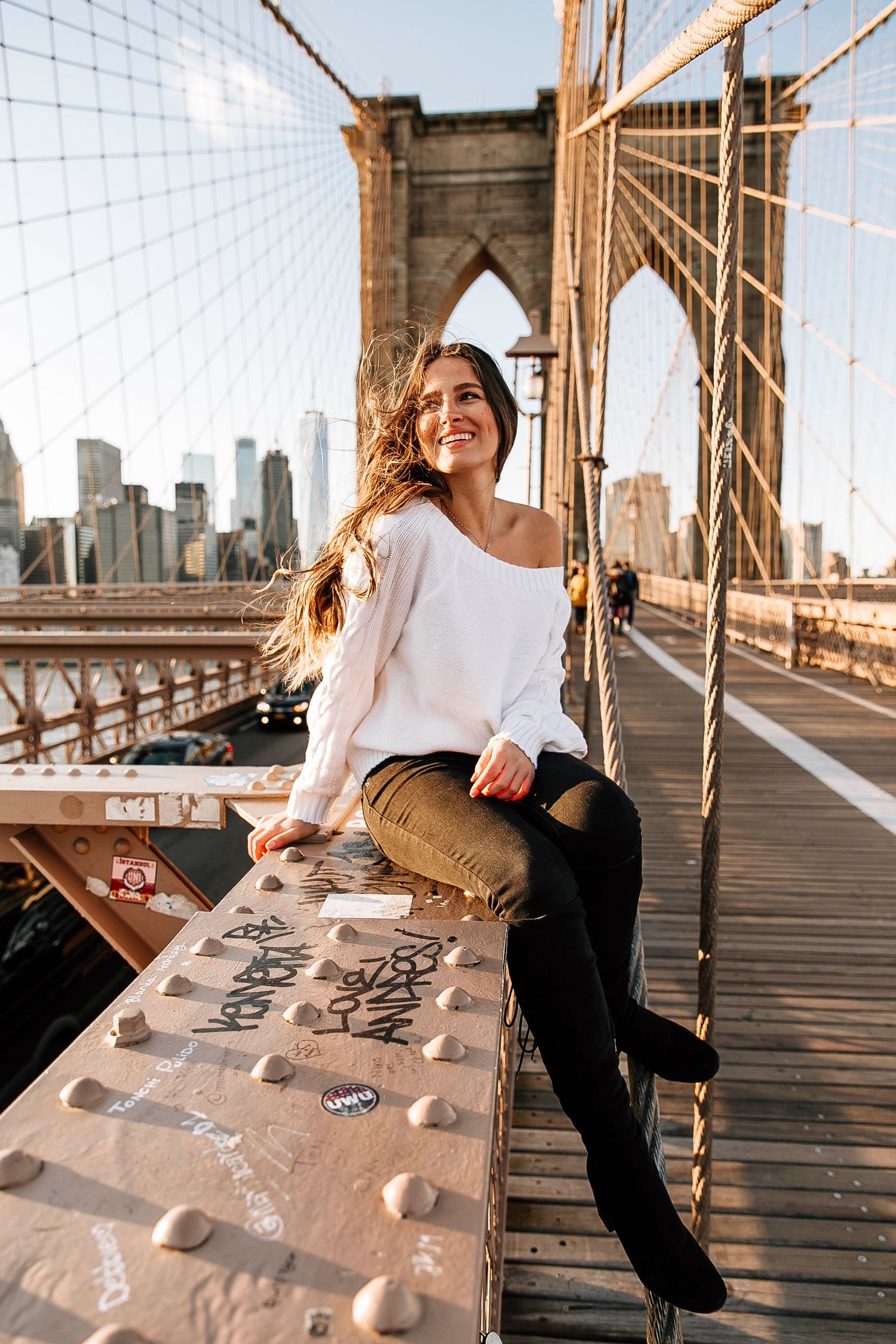 A woman in casual attire sits on the iconic Brooklyn Bridge during golden hour with the NYC skyline behind her.