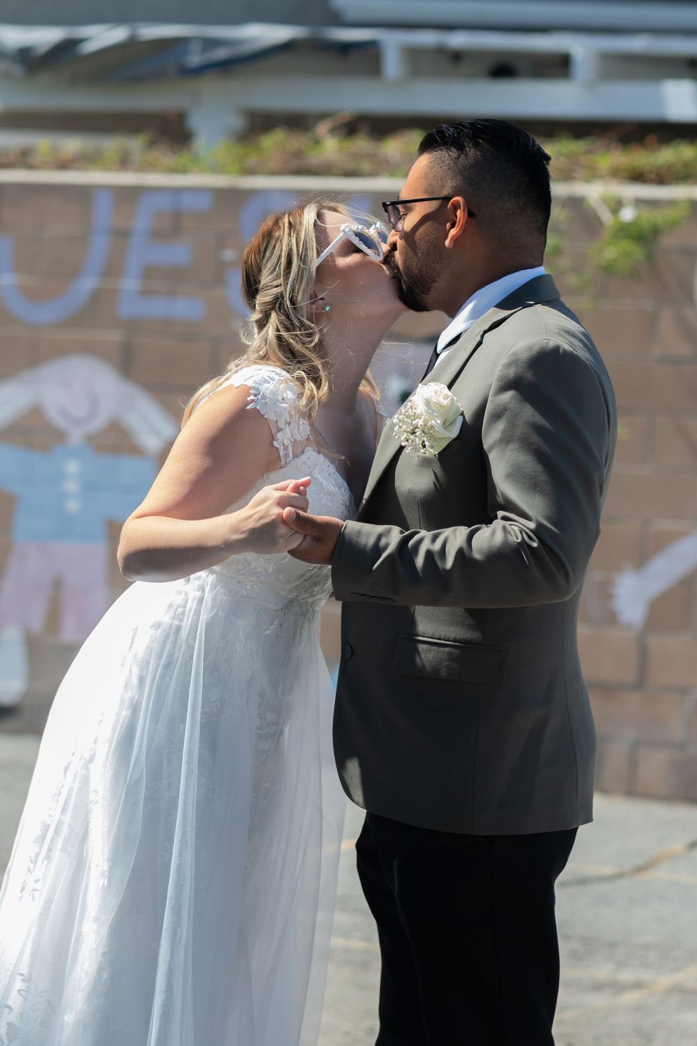Bride and groom sharing a kiss outdoors, bride in white lace gown, groom in gray suit with boutonniere.