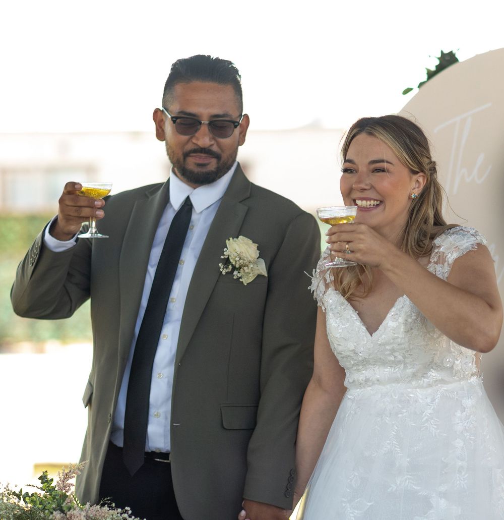 Bride and groom raising champagne glasses in a toast at their wedding ceremony, groom in olive suit.