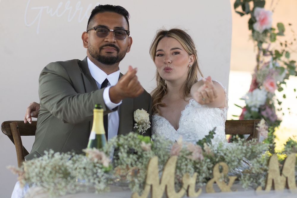 Bride and groom sitting at wedding reception table with champagne, floral arrangements, and Mr & Mrs sign.