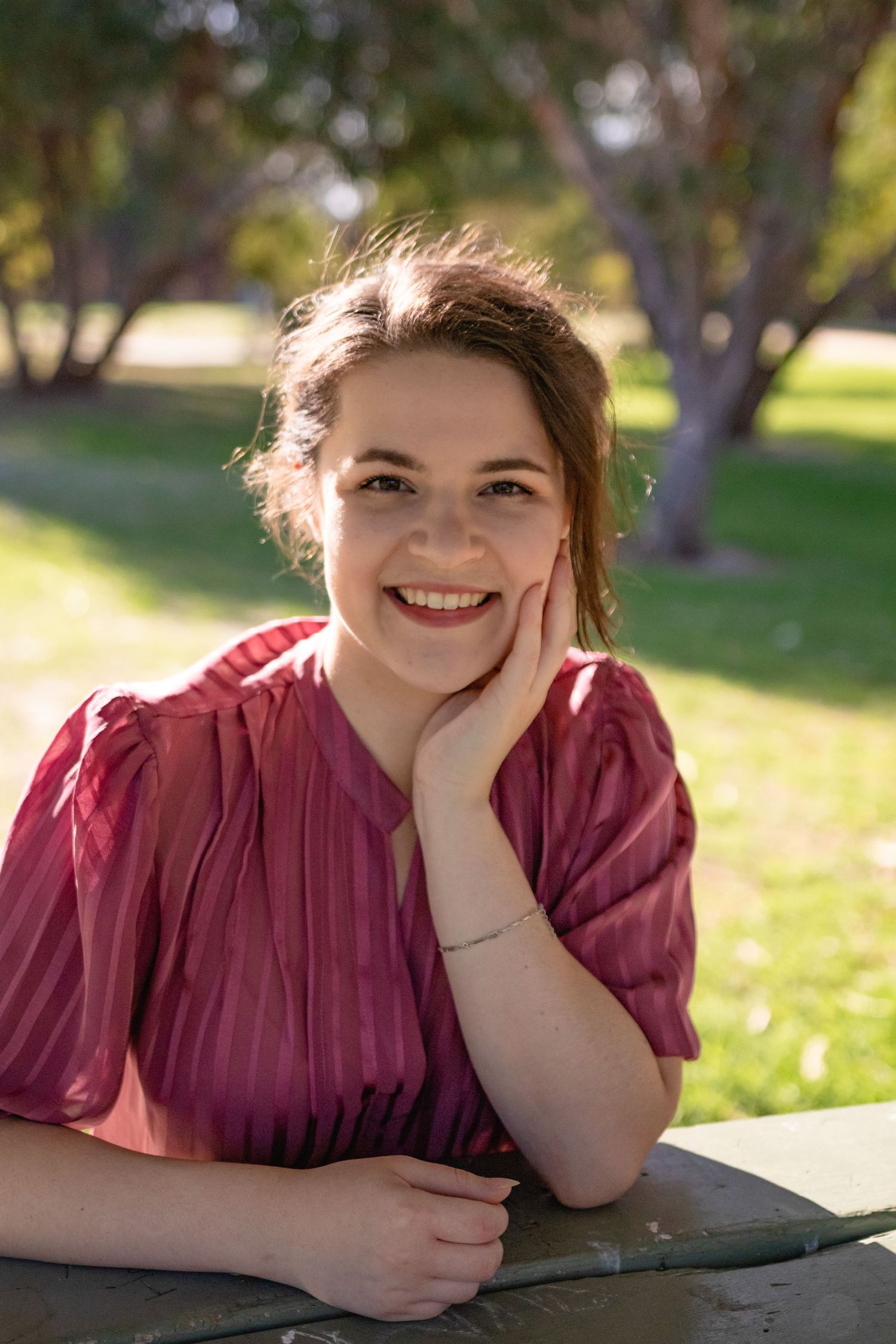 Smiling woman in a red blouse sits outdoors in a sunny Perth park, resting her chin on her hand during a branding session.