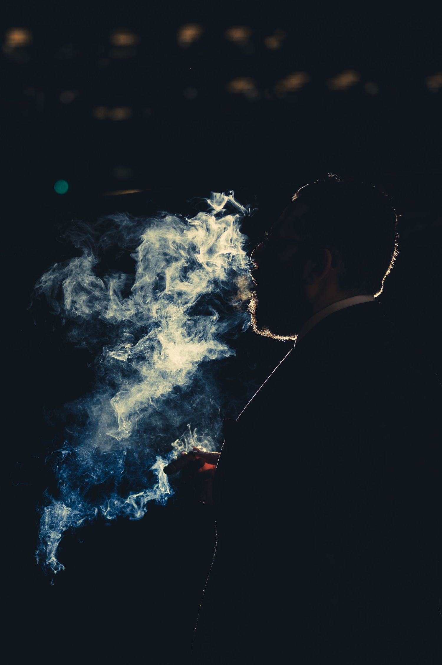 Man in gray suit smoking a cigar outdoors at night with dramatic smoke cloud behind him.